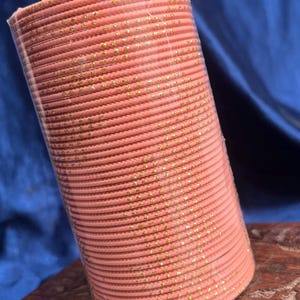 Op de afbeelding: Een stapel koraalkleurige armbanden met gouden vlekken, verpakt in doorzichtig plastic. De armbanden zijn netjes opgestapeld, waardoor een cilindrische vorm ontstaat. De achtergrond is een donkerblauwe stof.