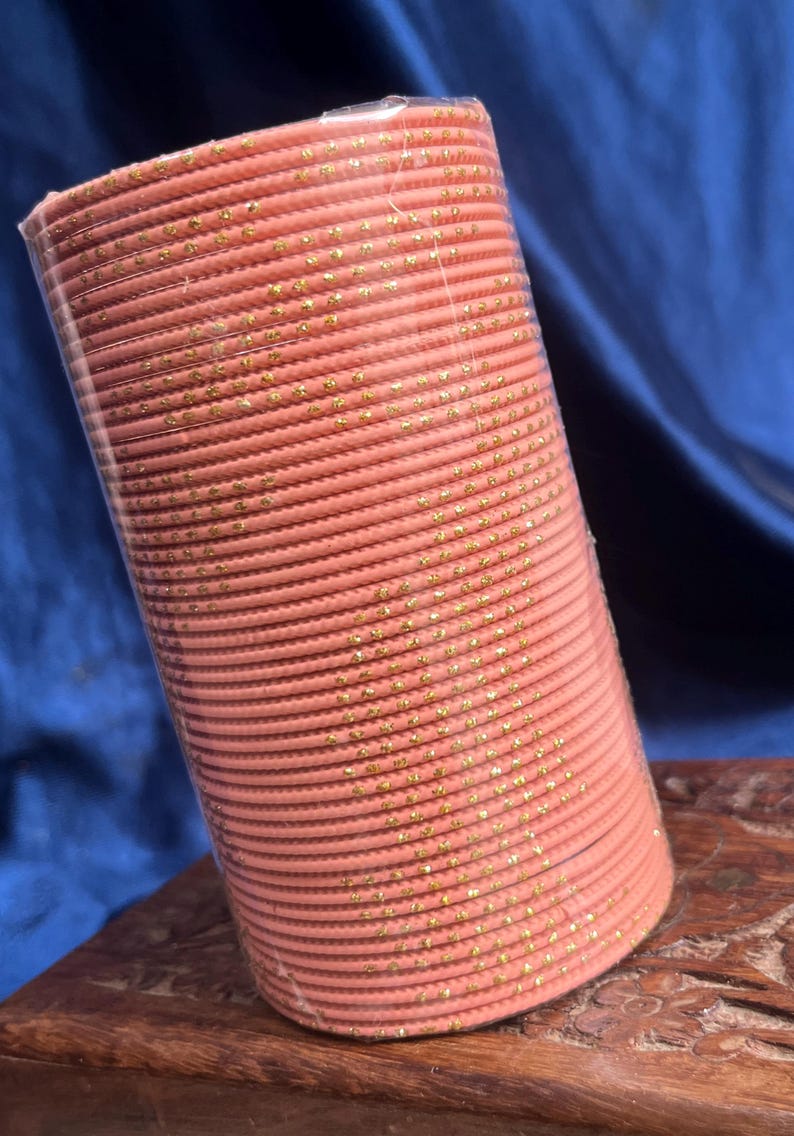 Op de afbeelding: Een stapel koraalkleurige armbanden met gouden vlekken, verpakt in doorzichtig plastic. De armbanden zijn netjes opgestapeld, waardoor een cilindrische vorm ontstaat. De achtergrond is een donkerblauwe stof.