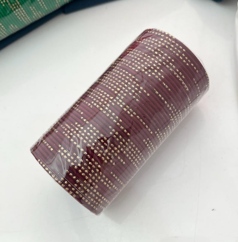 Op de afbeelding: Een bundel bordeauxrode armbanden met een gouden stippenpatroon, verpakt in doorzichtig plastic. De armbanden zijn op elkaar gestapeld, waardoor een cilindrische vorm ontstaat. De achtergrond is wit.