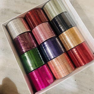 Op de afbeelding: Een doos gevuld met stapels kleurrijke armbanden. De armbanden zijn in verschillende kleuren, waaronder zilver, rood, roze, paars, groen en goud. De armbanden zijn gemaakt van metaal en hebben een glanzende afwerking. De doos is wit en de armbanden zijn netjes gerangschikt.