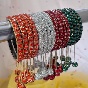 May include: A set of bangles featuring rows of orange, silver, red, and green rhinestones. Each bangle is adorned with matching dangling spherical charms suspended by thin gold-toned chains, creating a layered and decorative accessory set.