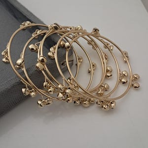 Può includere: Diversi braccialetti color oro con piccoli abbellimenti rotondi sono disposti su una superficie chiara. I braccialetti sono circolari e hanno una finitura lucida. Gli abbellimenti sono raggruppati lungo la circonferenza dei braccialetti.