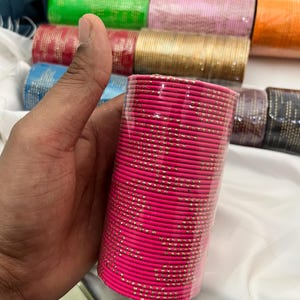 Op de afbeelding: Een stapel felroze armbanden met gouden accenten, vastgehouden in een hand. De armbanden zijn verpakt in doorzichtig plastic. Andere kleurrijke armbanden in groen, goud en roze zijn zichtbaar op de achtergrond.