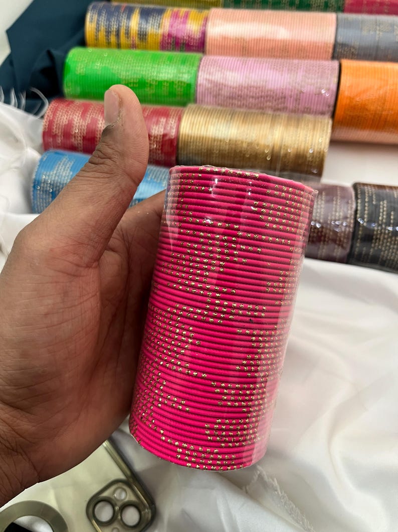 Op de afbeelding: Een stapel felroze armbanden met gouden accenten, vastgehouden in een hand. De armbanden zijn verpakt in doorzichtig plastic. Andere kleurrijke armbanden in groen, goud en roze zijn zichtbaar op de achtergrond.