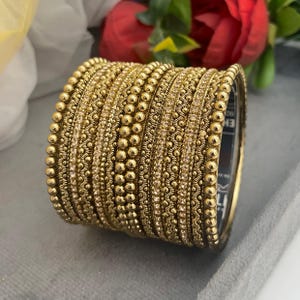 Könnte beinhalten: Ein Stapel goldener Armreifen mit aufwendigen Designs. Die Armreifen weisen eine Mischung aus Texturen auf, darunter kleine Perlen und detaillierte Muster. Einige Armreifen haben eine Reihe kleiner, klarer Steine. Der Schmuck wird auf einer grauen Oberfläche präsentiert.