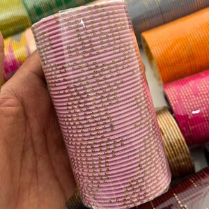 Op de afbeelding: Een bundel roze armbanden met gouden accenten, verpakt in doorzichtig plastic. Andere kleurrijke armbanden zijn zichtbaar op de achtergrond. De armbanden zijn waarschijnlijk van metaal.