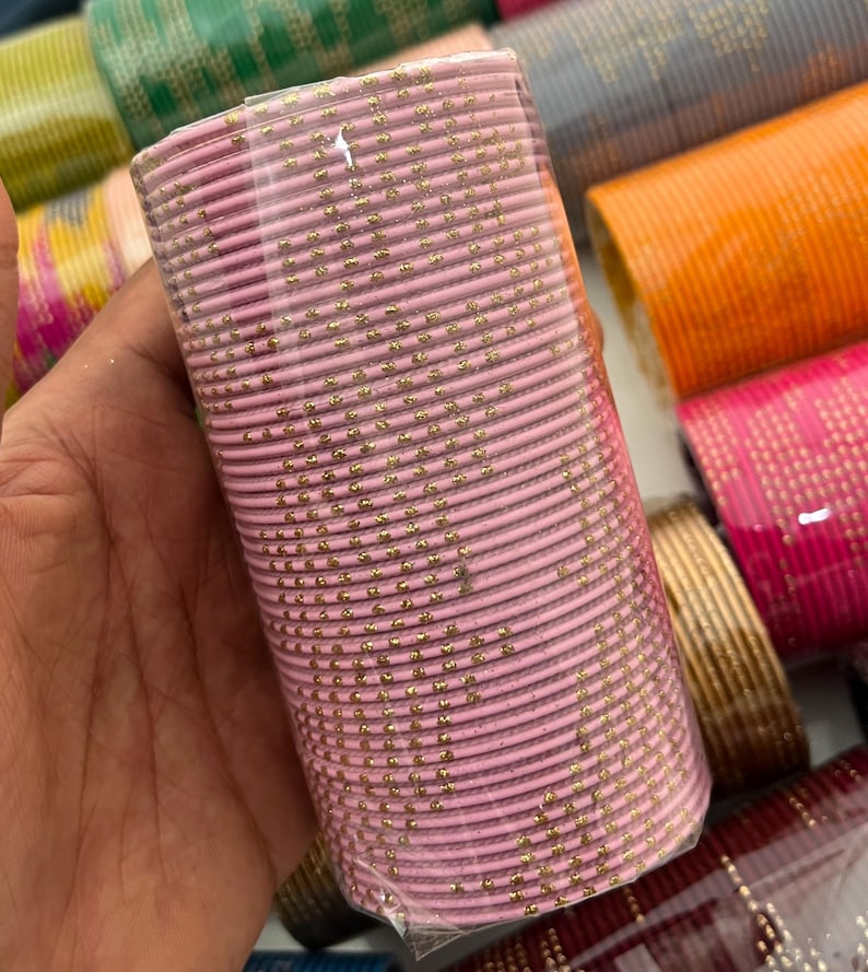 Op de afbeelding: Een bundel roze armbanden met gouden accenten, verpakt in doorzichtig plastic. Andere kleurrijke armbanden zijn zichtbaar op de achtergrond. De armbanden zijn waarschijnlijk van metaal.