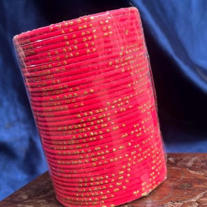 Op de afbeelding: Een stapel rode armbanden met gouden stippenpatronen, verpakt in doorzichtig plastic. De armbanden zijn gestapeld op een gebeeldhouwd houten oppervlak, met een donkerblauwe achtergrond.
