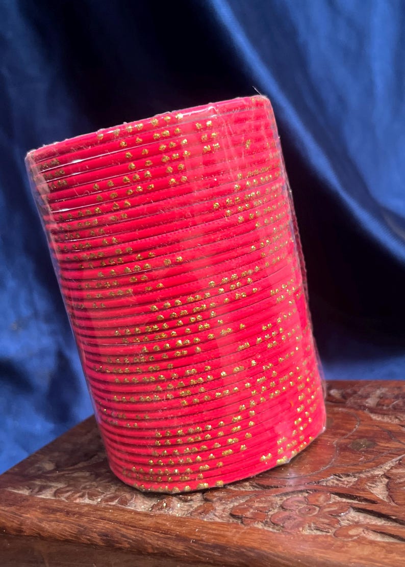 Op de afbeelding: Een stapel rode armbanden met gouden stippenpatronen, verpakt in doorzichtig plastic. De armbanden zijn gestapeld op een gebeeldhouwd houten oppervlak, met een donkerblauwe achtergrond.