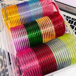 Puede incluir: Un contenedor de almacenamiento de plástico transparente lleno de numerosos brazaletes finos, redondos y translúcidos en una variedad de colores vibrantes que incluyen amarillo, azul, rojo, verde, rosa y lima, dispuestos en filas ordenadas.