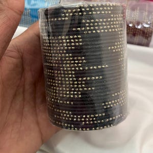Op de afbeelding: Een stapel zwarte armbanden met goudkleurige versieringen, verpakt in doorzichtig plastic. De armbanden zijn cilindrisch en lijken van een stevig materiaal te zijn gemaakt. Andere stapels armbanden zijn op de achtergrond zichtbaar.