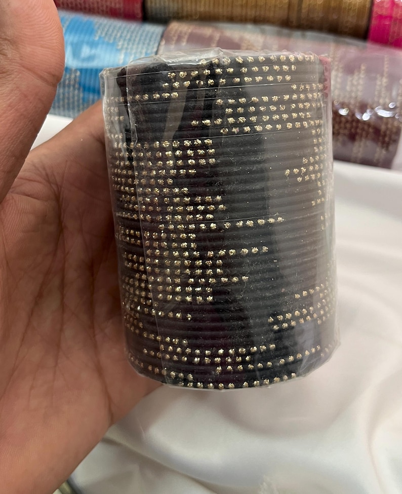 Op de afbeelding: Een stapel zwarte armbanden met goudkleurige versieringen, verpakt in doorzichtig plastic. De armbanden zijn cilindrisch en lijken van een stevig materiaal te zijn gemaakt. Andere stapels armbanden zijn op de achtergrond zichtbaar.