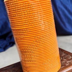 Op de afbeelding: Een stapel oranje armbanden met gouden accenten. De armbanden zijn cilindrisch en lijken van metaal of een vergelijkbaar materiaal te zijn. De stapel is verpakt in doorzichtig plastic, de achtergrond is een donkerblauwe stof en een houten oppervlak.