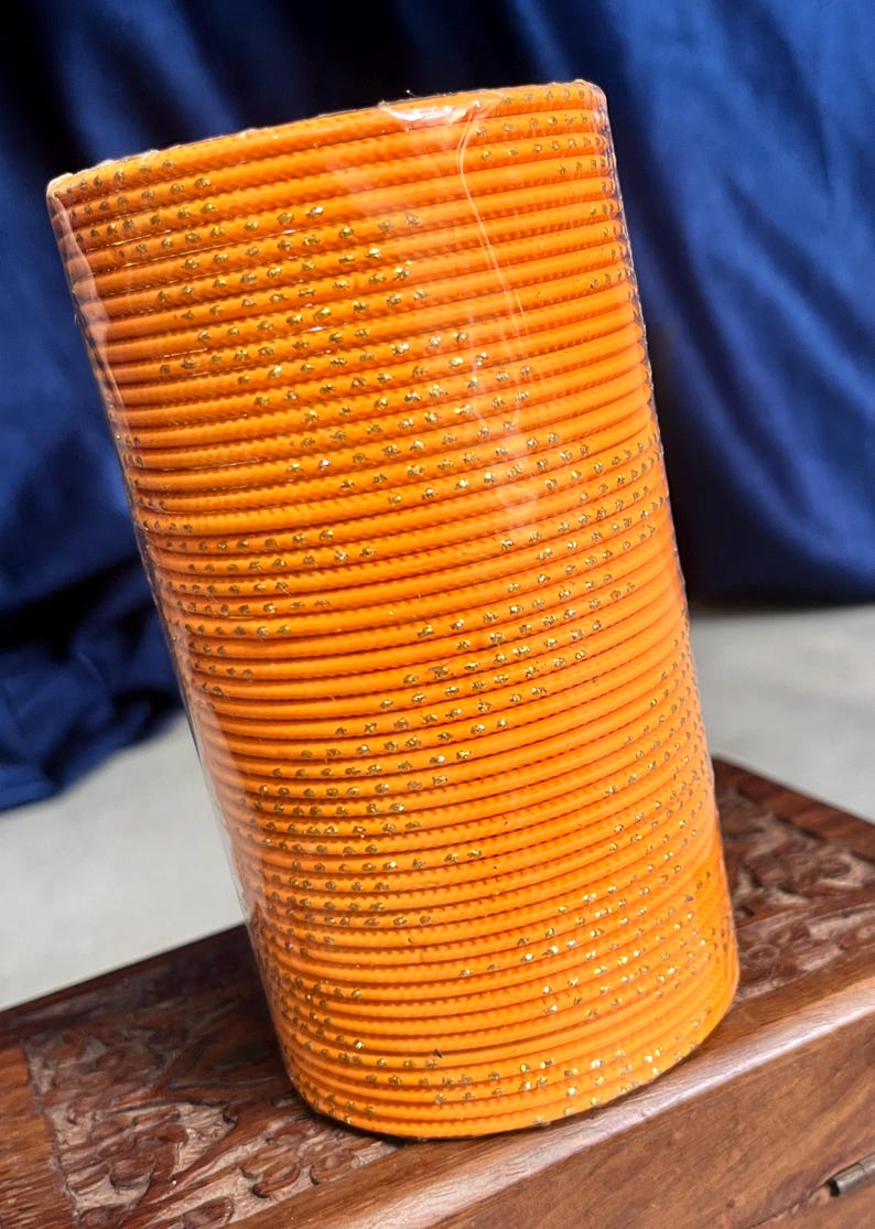 Op de afbeelding: Een stapel oranje armbanden met gouden accenten. De armbanden zijn cilindrisch en lijken van metaal of een vergelijkbaar materiaal te zijn. De stapel is verpakt in doorzichtig plastic, de achtergrond is een donkerblauwe stof en een houten oppervlak.