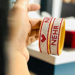 Puede incluir: Dos pulseras amarillas con detalles rojos y el nombre "NEHA" en letras rojas. Las pulseras están adornadas con pequeños y brillantes pedrería. Un corazón rojo también aparece en la pulsera.