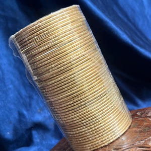 Op de afbeelding: Een bundel goudkleurige armbanden, strak verpakt in doorzichtig plastic. De armbanden zijn in een cilindrische vorm gestapeld, met een lichte glans. De achtergrond is een diepblauwe stof en de armbanden rusten op een houten oppervlak.