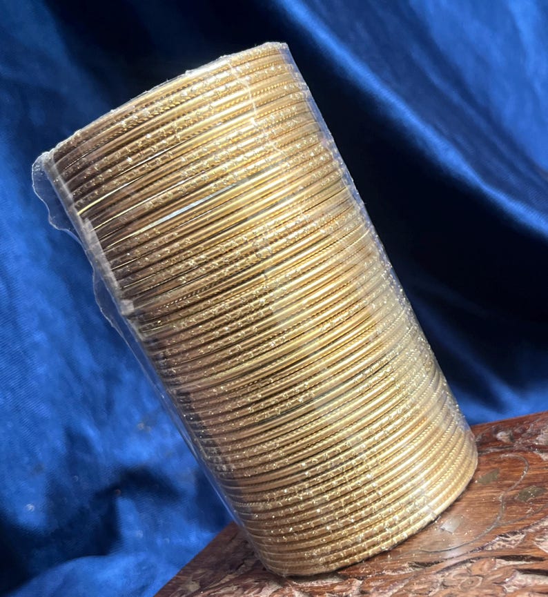 Op de afbeelding: Een bundel goudkleurige armbanden, strak verpakt in doorzichtig plastic. De armbanden zijn in een cilindrische vorm gestapeld, met een lichte glans. De achtergrond is een diepblauwe stof en de armbanden rusten op een houten oppervlak.