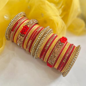 May include: A stack of bangles featuring alternating red and gold bands. The set includes textured gold bands, smooth red bands, and decorative bands encrusted with small white pearls and red floral accents arranged in a cylindrical formation.
