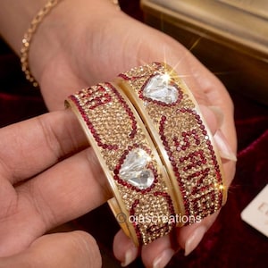 Puede incluir: Dos pulseras doradas adornadas con cristales rojos y transparentes. Cada pulsera presenta un cristal en forma de corazón y la palabra "LOVE" en cristales rojos. Una pulsera dorada es visible en el fondo.