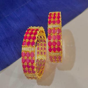 May include: Two gold-toned bangles with vibrant pink oval-cut gemstones and rectangular clear stones. The bangles are set against a blue fabric background, showcasing the jewelry's intricate design and color contrast.