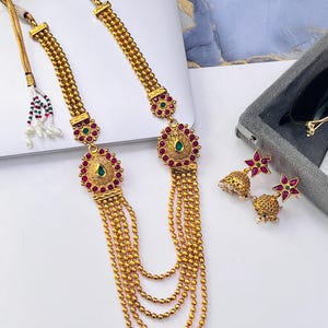 May include: A gold-toned necklace featuring multiple beaded chains, circular pendants with pink and green stones, and matching jhumka earrings with star-shaped tops. The necklace includes an adjustable cord with white and green beaded tassels.