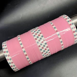May include: A stack of pink bangles with white bands featuring a pattern of red, blue, and black circles. The bangles are cylindrical and appear to be made of a glossy material. The background is dark.