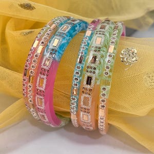 Puede incluir: Un conjunto de pulseras coloridas. Las pulseras presentan un degradado de colores, incluyendo rosa, azul y verde. Cada pulsera está adornada con adornos rectangulares y redondos. El fondo es una tela amarilla.