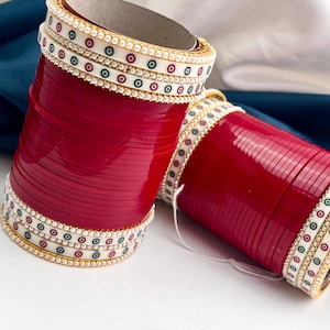 May include: Two red bangles with white and gold detailing. The bangles feature small, colourful dots and faux pearls. The design is repeated around the circumference. The bangles are stacked together, showing the intricate design.