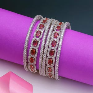 May include: A set of silver bangles featuring white and red gemstones. The bangles are displayed on a purple surface. The gemstones are rectangular and set in a detailed pattern. The overall design is elegant and eye-catching.