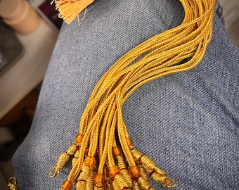 Puede incluir: Un conjunto de borlas de cordón amarillo dorado con un nudo superior trenzado y detalles en rojo oscuro. Cada cordón termina en una sección de hilo dorado envuelto, una pequeña cuenta de color ámbar y un pequeño lazo de metal dorado.