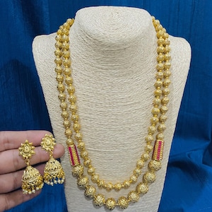 Pode incluir: Um colar dourado com duas fileiras de contas, exibido em um manequim. O colar apresenta contas redondas e texturizadas. Brincos combinando com um design floral e pérolas penduradas também são visíveis.