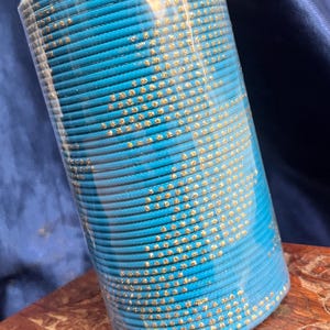 Op de afbeelding: Een stapel turquoise armbanden met goudkleurige versieringen. De armbanden zijn verpakt in helder plastic en liggen op een houten oppervlak. De gouden accenten contrasteren met het blauw.