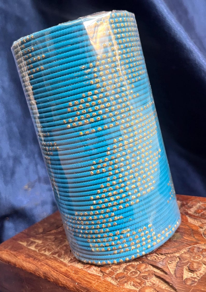 Op de afbeelding: Een stapel turquoise armbanden met goudkleurige versieringen. De armbanden zijn verpakt in helder plastic en liggen op een houten oppervlak. De gouden accenten contrasteren met het blauw.