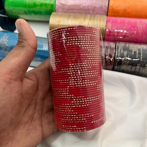 Op de afbeelding: Een bundel rode armbanden met kleine goudkleurige versieringen. De armbanden zijn verpakt in doorzichtig plastic. Op de achtergrond zijn andere bundels armbanden in verschillende kleuren te zien, waaronder roze, groen en goud.