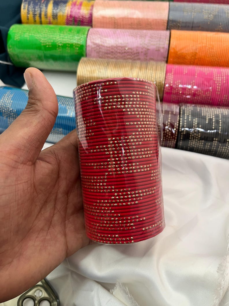 Op de afbeelding: Een bundel rode armbanden met kleine goudkleurige versieringen. De armbanden zijn verpakt in doorzichtig plastic. Op de achtergrond zijn andere bundels armbanden in verschillende kleuren te zien, waaronder roze, groen en goud.