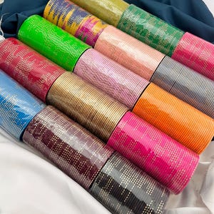 Könnte beinhalten: Eine Sammlung farbenfroher Armreifen in verschiedenen Farbtönen, darunter Grün, Rosa, Gold und Blau. Jeder Armreif ist mit einem gepunkteten Muster verziert. Die Armreifen sind in Reihen angeordnet und bilden eine ansprechende Präsentation von Accessoires.