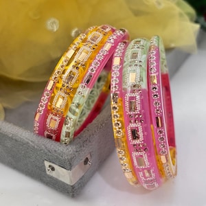 Puede incluir: Un conjunto de pulseras coloridas en tonos rosa, amarillo y verde. Las pulseras están adornadas con elementos rectangulares y circulares. Se exhiben sobre una superficie de terciopelo gris. Las pulseras son un accesorio atractivo.