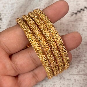 Puede incluir: Un conjunto de cuatro brazaletes dorados con un patrón ondulado repetitivo y detalles texturizados complejos en la superficie. Las pulseras circulares tienen un acabado metálico pulido y un ancho constante, mostrando un diseño en relieve.