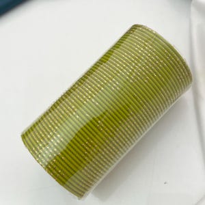 Op de afbeelding: Een stapel olijfgroene armbanden met gouden accenten. De cilindrische armbanden zijn op elkaar gestapeld en verpakt in helder plastic. De armbanden zijn waarschijnlijk gemaakt van een stevig materiaal.