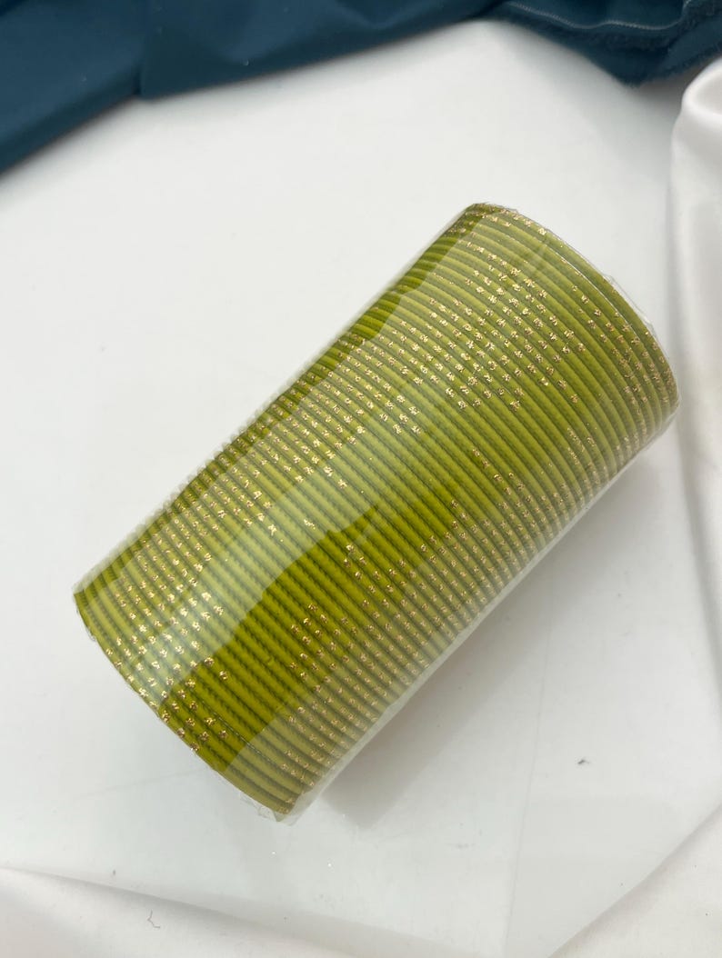 Op de afbeelding: Een stapel olijfgroene armbanden met gouden accenten. De cilindrische armbanden zijn op elkaar gestapeld en verpakt in helder plastic. De armbanden zijn waarschijnlijk gemaakt van een stevig materiaal.