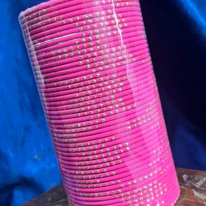 Op de afbeelding: Een stapel felroze armbanden met goudkleurige versieringen. De armbanden zijn cilindrisch en verpakt in helder plastic. De achtergrond is een donkerblauwe stof en de armbanden liggen op een houten oppervlak.