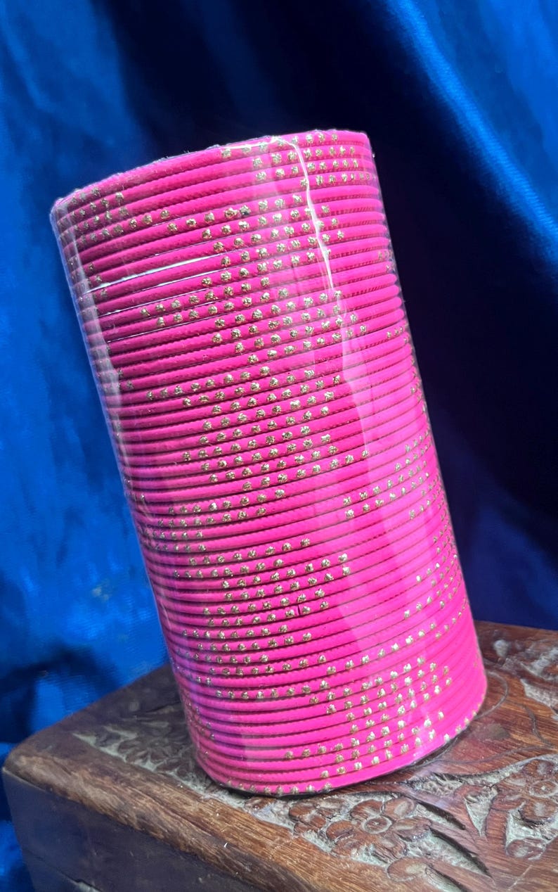 Op de afbeelding: Een stapel felroze armbanden met goudkleurige versieringen. De armbanden zijn cilindrisch en verpakt in helder plastic. De achtergrond is een donkerblauwe stof en de armbanden liggen op een houten oppervlak.