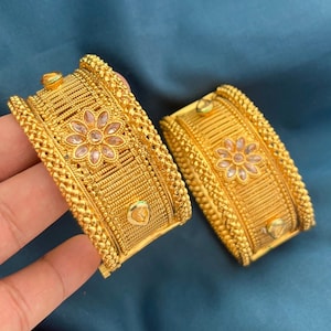 May include: Two gold-colored bangles with intricate detailing. Each bangle features a central floral design with pink gemstones and is adorned with textured patterns and small embellishments. The bangles are displayed against a blue background.