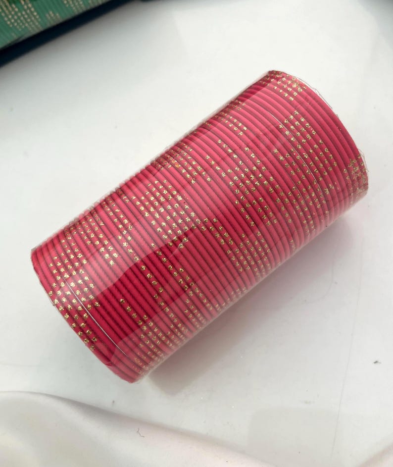 Op de afbeelding: Een stapel roze armbanden met gouden accenten. De armbanden zijn verpakt in doorzichtig plastic. De gouden accenten zijn kleine, gelijkmatig verdeelde stippen. De armbanden zijn een levendige roze kleur.