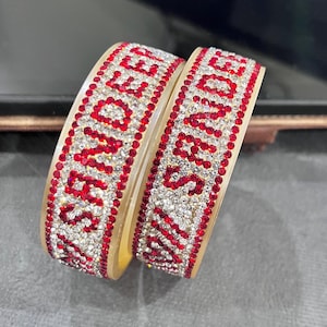 May include: Two ivory bangles decorated with red and clear crystals. The bangles feature a word spelled out in red crystals. The edges are lined with small red crystals. The bangles are displayed on a dark surface.