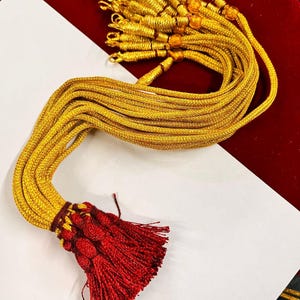 May include: A collection of gold-colored cords with small loops at the top, bundled together. The cords transition to a dark red tassel with a braided section. The background is a mix of red and white.