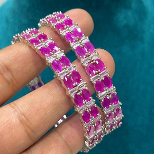 May include: Two silver bangles, each featuring alternating rows of oval-cut fuchsia gemstones and rectangular clear stones. The bangles are displayed against a teal background, highlighting the jewellery's vibrant colours and design.
