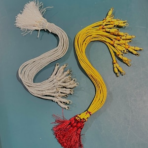 Pode incluir: Duas borlas decorativas. Uma é prateada com fechos prateados, e a outra é amarela com fechos dourados e uma borla vermelha na extremidade. As borlas são feitas de um material tecido e provavelmente são usadas para embelezar.