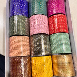 May include: A box filled with stacks of colorful bangles. The bangles are in various colors, including blue, green, pink, red, peach, black, and gold. Each bangle is adorned with small, gold-colored embellishments.