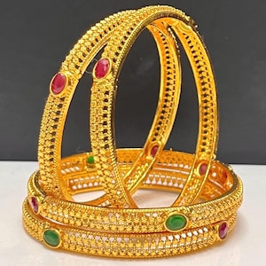 May include: Two gold bangles featuring a textured, intricate lattice pattern along the outer band. Each bangle is accented with alternating oval-shaped red and green stones set at regular intervals along the circumference of the metal surface.